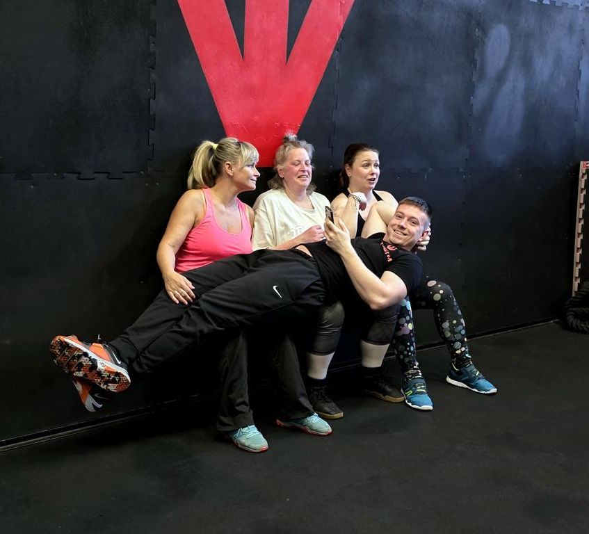 Tři klientky FitWay40 s trenérem se smějí při společném cviku u zdi